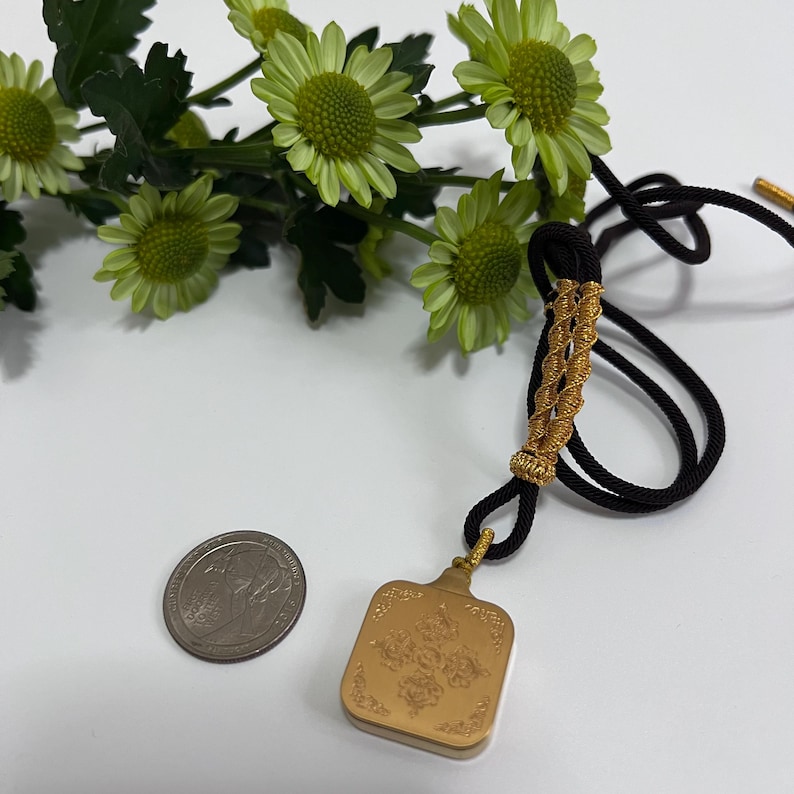 May include: A square gold-colored pendant with an embossed floral pattern, attached to a black cord with gold-wrapped accents. A silver-colored US quarter coin is placed next to the pendant for scale, and green chrysanthemum flowers are visible.
