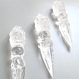 May include: Three clear, transparent quartz crystal carvings shaped like phurba daggers. Each piece features a detailed, sculpted head at the top handle, transitioning into a smooth, pointed blade with intricate, carved geometric patterns.