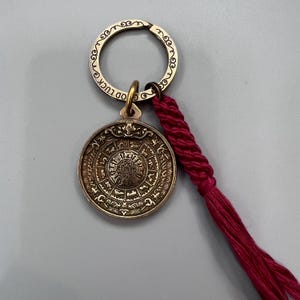 Puede incluir: Un llavero dorado con un dije redondo que presenta intrincados caracteres chinos y una borla roja. El dije tiene las palabras "Good Luck" inscritas en el anillo.