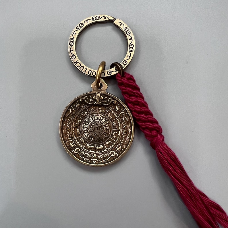 Puede incluir: Un llavero dorado con un dije redondo que presenta intrincados caracteres chinos y una borla roja. El dije tiene las palabras "Good Luck" inscritas en el anillo.