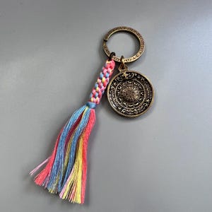 Puede incluir: Un llavero dorado con un dije redondo que presenta un dise&ntilde;o circular con detalles intrincados. El dije est&aacute; unido a un llavero con una borla trenzada de color rosa, azul y amarillo.