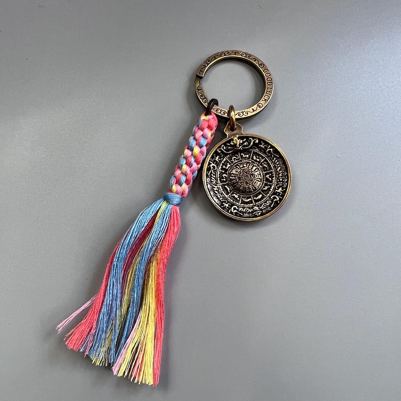 Puede incluir: Un llavero dorado con un dije redondo que presenta un dise&ntilde;o circular con detalles intrincados. El dije est&aacute; unido a un llavero con una borla trenzada de color rosa, azul y amarillo.