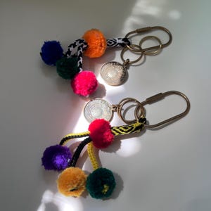 Puede incluir: Dos llaveros con anillos ovalados y dijes circulares en tono latón. Cada llavero tiene cordones trenzados adornados con pompones multicolores en tonos naranja, azul, verde, amarillo y rosa, sujetos a los herrajes metálicos.