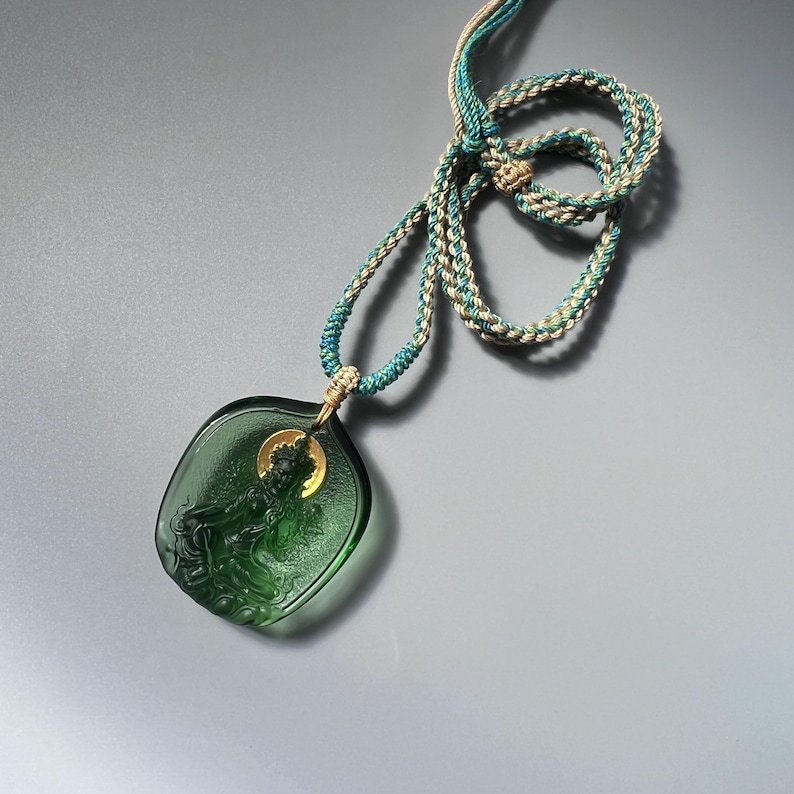 May include: A green glass pendant necklace with a detailed carving of a figure. The necklace is made with a braided cord in shades of teal and beige.