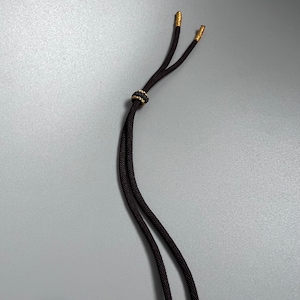 May include: A black cord with a braided texture featuring two gold-toned metal tips at the ends. A small, round, black and gold-toned decorative bead is positioned on the cord, allowing for adjustment of the two parallel strands of the cord.