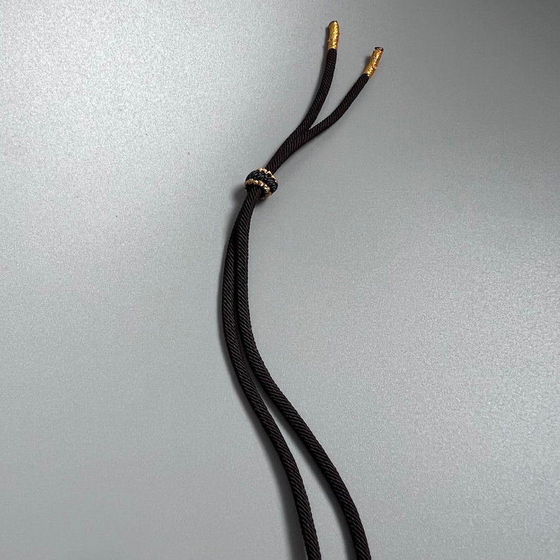 May include: A black cord with a braided texture featuring two gold-toned metal tips at the ends. A small, round, black and gold-toned decorative bead is positioned on the cord, allowing for adjustment of the two parallel strands of the cord.