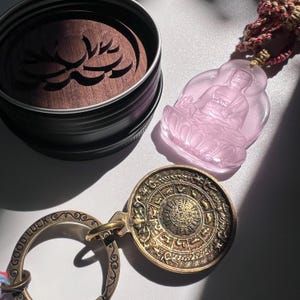 May include: A collection of spiritual items, including a pink Buddha pendant, a bronze-coloured keychain with the words "GOOD LUCK", and a round, engraved pendant. A dark metal container with a wooden lid featuring a carved design is also present.