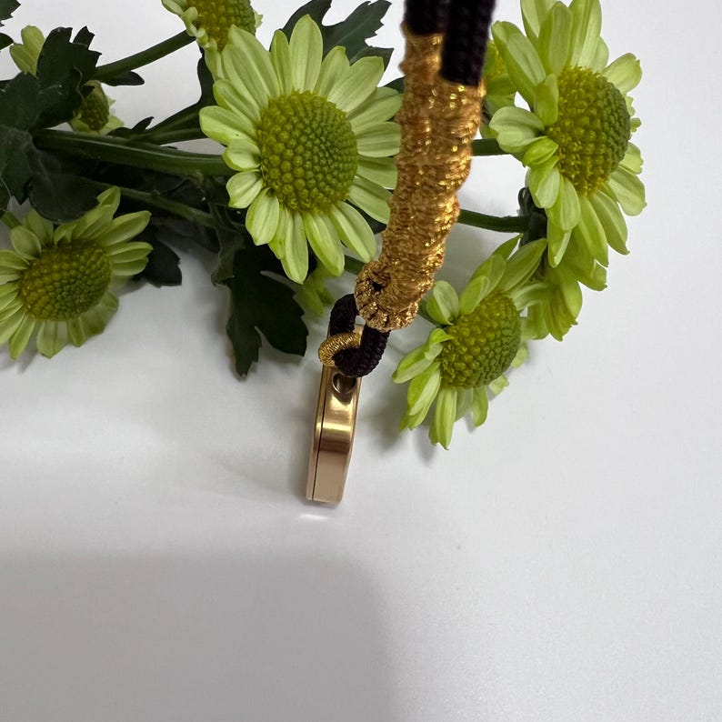 May include: A gold-toned metal pendant with a smooth, rectangular profile, suspended from a dark brown cord wrapped in textured gold thread. The pendant is positioned against a cluster of bright green chrysanthemum flowers with yellow centers.