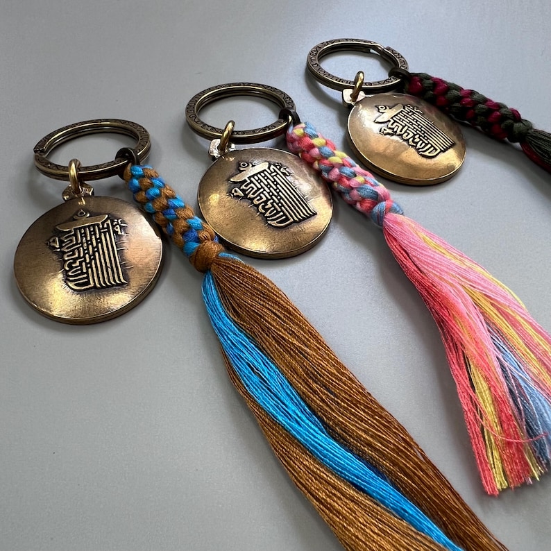 Puede incluir: Cuatro llaveros con dijes redondos de metal que presentan caracteres chinos y borlas trenzadas de colores. Las borlas son marr&oacute;n y azul, rosa y amarillo, rosa y amarillo y granate y verde.