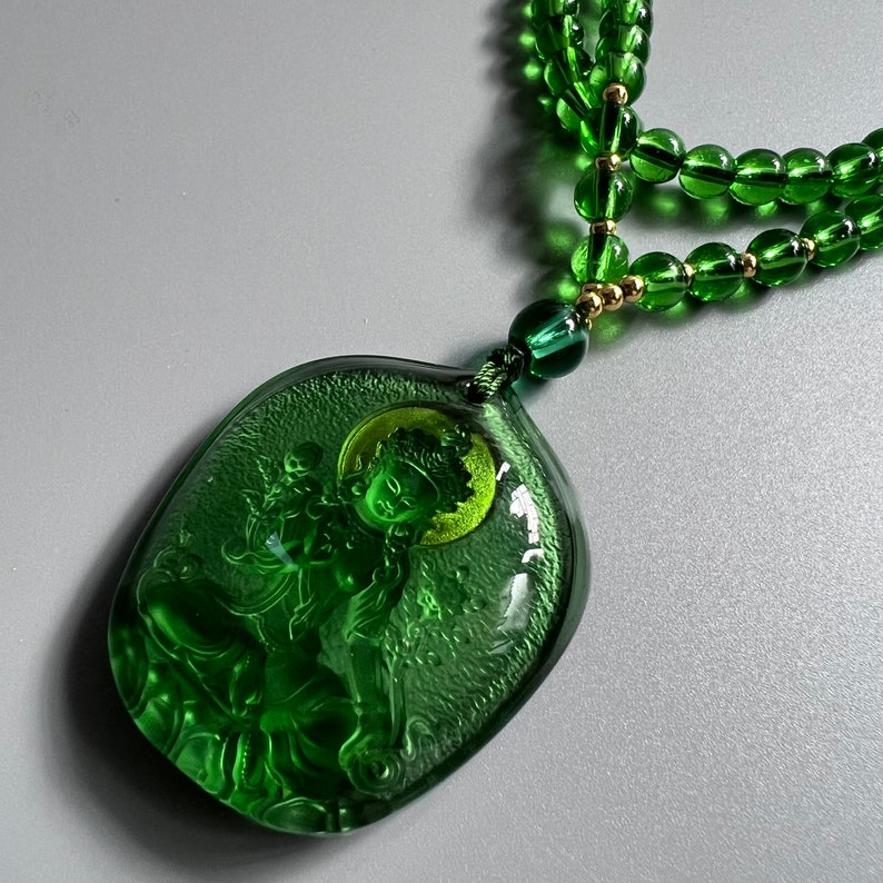 May include: A green jade necklace with a carved pendant featuring a figure of a deity. The pendant is intricately detailed and has a green and gold color scheme.