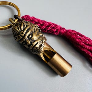 Puede incluir: Un llavero de silbato de latón con un rostro esculpido con barba y una superficie texturizada. El silbato está unido a una anilla circular de latón y decorado con una larga borla de cordón de color magenta vibrante en la parte superior.