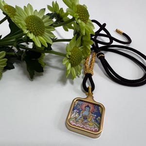 May include: A square pendant featuring a colorful illustration of a seated figure in a gold-toned frame, suspended from a black cord with gold-colored decorative accents. The pendant is positioned next to a cluster of light green chrysanthemum flowers.