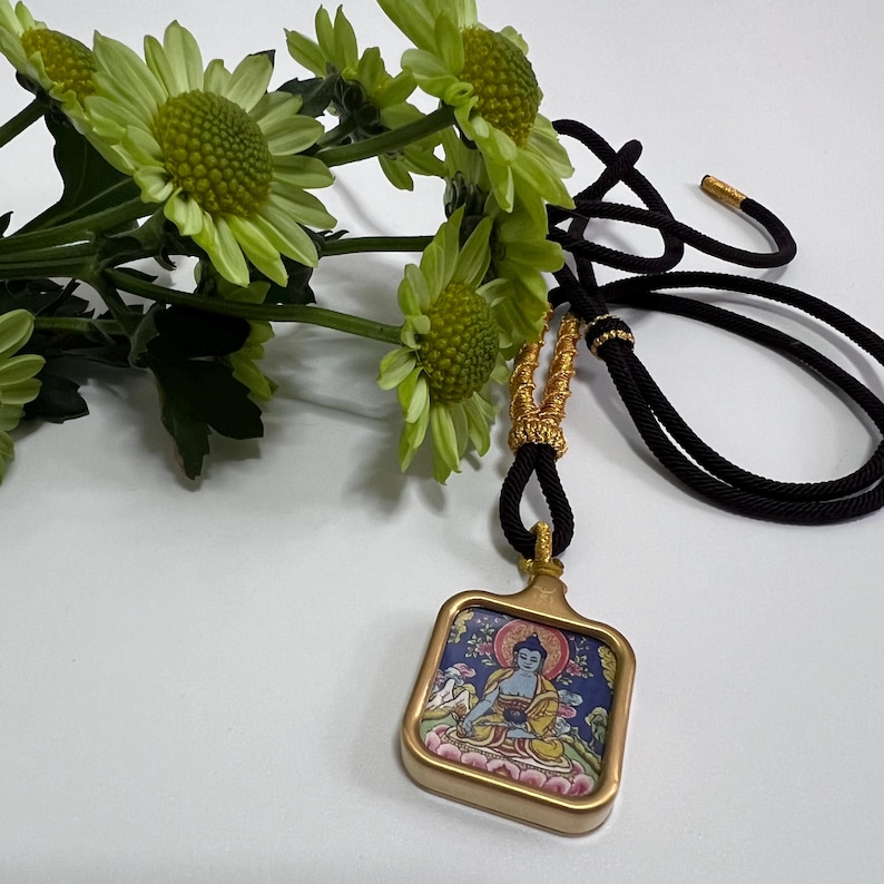 May include: A square pendant featuring a colorful illustration of a seated figure in a gold-toned frame, suspended from a black cord with gold-colored decorative accents. The pendant is positioned next to a cluster of light green chrysanthemum flowers.