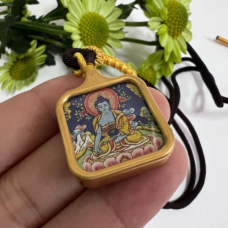 May include: A square pendant featuring a colorful illustration of a seated figure in yellow and blue robes against a dark blue background, encased in a matte gold frame with a dark brown cord and a gold decorative knot at the top.