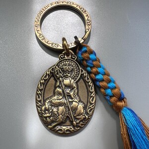 Puede incluir: Un llavero de metal en tono bronce con una anilla circular grabada con las palabras GOOD LUCK. Incluye un colgante ovalado que representa una figura sentada sosteniendo un bastón, junto a un cordón trenzado azul y marrón con una borla.