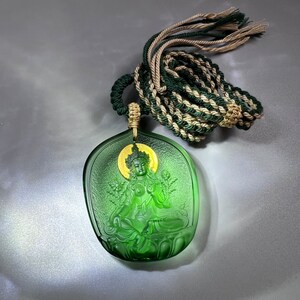 May include: A green glass pendant with a detailed carving of a seated figure. The pendant is attached to a green and beige braided cord.