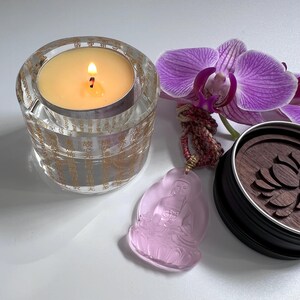 May include: A lit candle in a clear glass votive with gold text, a pink Buddha pendant, a purple orchid, and a wooden coaster with a lotus design. The candle flame glows brightly, casting a warm light.