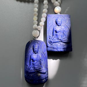 May include: Two blue lapis lazuli pendants with carved Buddha figures, suspended on beaded necklaces. The pendants are rectangular and oval, with the Buddha in a seated pose. The necklaces have white beads and a pearlescent clasp.