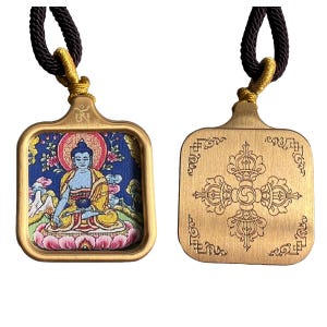 May include: Two square brass pendants on dark brown braided cords. One pendant features a colorful fabric inlay of a seated blue figure on a pink lotus, with a gold Om symbol above. The other pendant displays an engraved cross-shaped vajra design.