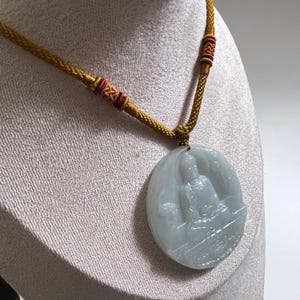 May include: A pale green jade pendant necklace with a carved seated figure in an oval. The pendant is suspended from a gold-coloured woven cord with red and gold details. The necklace is displayed on a mannequin.