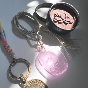 Puede incluir: Un collar con colgante rosa con una figura tallada, un cordón trenzado y una borla. También hay un pequeño recipiente redondo negro con una tapa de madera con un diseño tallado. También es visible un llavero con las palabras "GOOD LUCK".