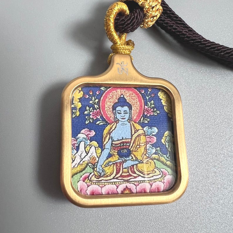 May include: A rectangular pendant with a brushed gold-tone metal frame featuring a central image of a blue-skinned figure in a yellow robe seated on a pink lotus. The pendant is suspended by a dark brown cord with gold-tone decorative knotting.