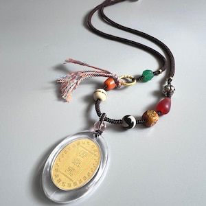 May include: A gold pendant necklace with an oval pendant in clear plastic. The pendant has Chinese characters. The necklace features a dark brown cord, colourful beads, and a tassel.