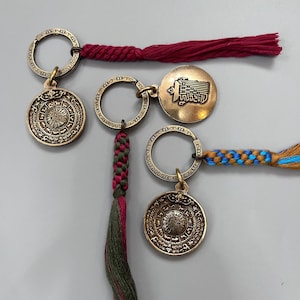 Puede incluir: Tres llaveros de color dorado con dise&ntilde;os intrincados. Cada llavero presenta un dije circular con s&iacute;mbolos grabados y un peque&ntilde;o anillo. Los llaveros est&aacute;n unidos a cordones trenzados de colores con borlas.