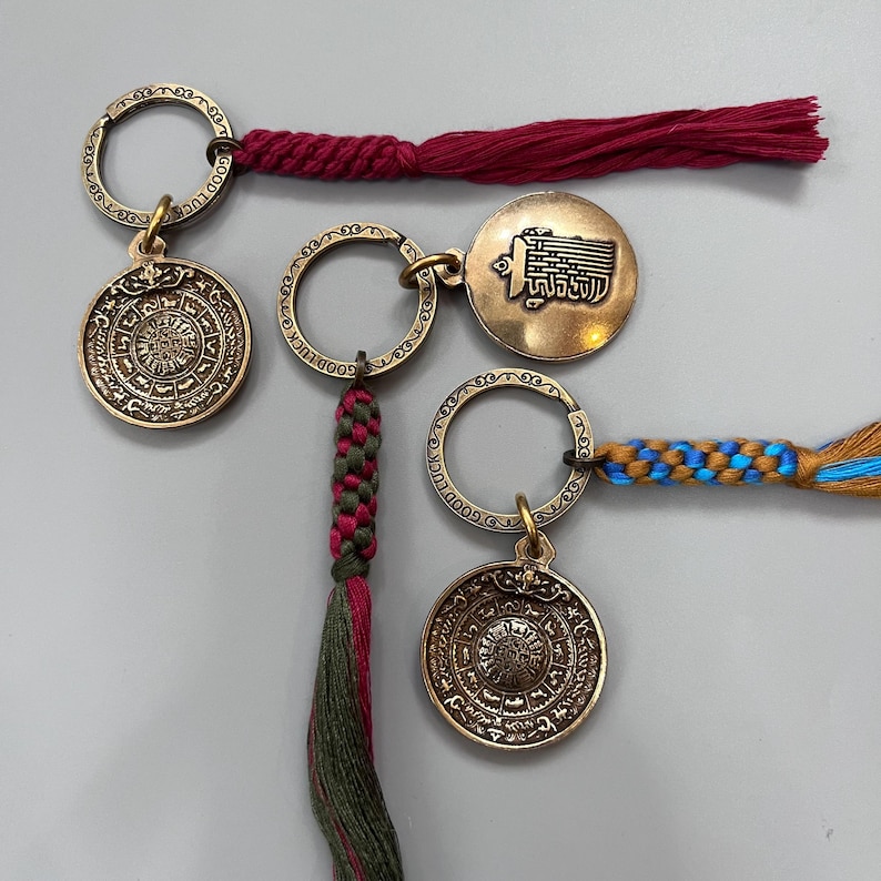 Puede incluir: Tres llaveros de color dorado con dise&ntilde;os intrincados. Cada llavero presenta un dije circular con s&iacute;mbolos grabados y un peque&ntilde;o anillo. Los llaveros est&aacute;n unidos a cordones trenzados de colores con borlas.