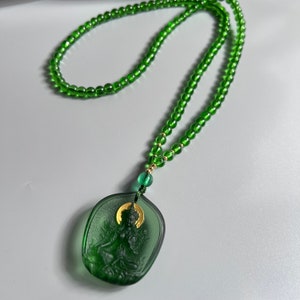 May include: A green glass necklace with a green glass pendant featuring a carved figure. The pendant has a gold accent.