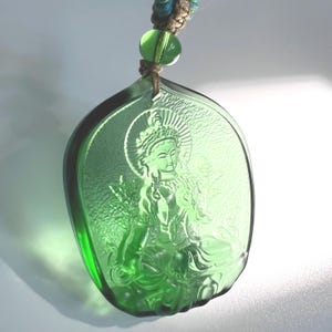 Pode incluir: Um pingente verde translúcido com uma imagem gravada de uma figura sentada com um halo. O pingente em forma de folha é suspenso por um cordão com uma conta verde.