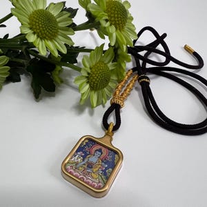 May include: A square pendant featuring a colorful illustration of a seated figure in a gold-toned frame, suspended from a black cord with gold-toned accents. The pendant is positioned near a cluster of light green chrysanthemum flowers.