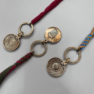 Puede incluir: Dos llaveros de oro con dise&ntilde;os intrincados y cordones trenzados rojos, verdes y azules. Un llavero presenta un dije redondo con escritura &aacute;rabe y el otro presenta un dije redondo con una imagen estilizada de un edificio.