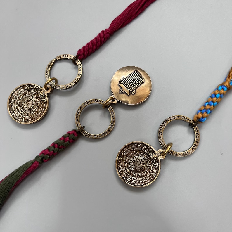 Puede incluir: Dos llaveros de oro con dise&ntilde;os intrincados y cordones trenzados rojos, verdes y azules. Un llavero presenta un dije redondo con escritura &aacute;rabe y el otro presenta un dije redondo con una imagen estilizada de un edificio.