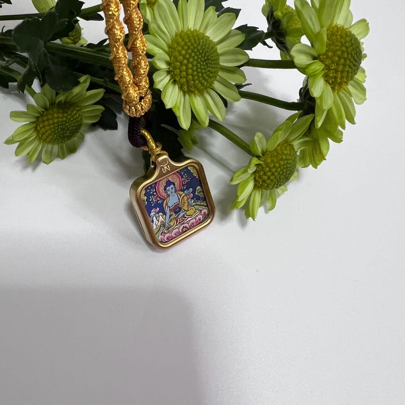 May include: A square gold-toned pendant featuring a colorful miniature painting of a seated figure in blue and yellow robes on a dark blue background, suspended from a thick, braided gold-colored cord with a dark brown accent near the top.