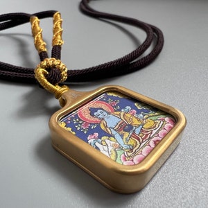 May include: A rectangular pendant with a matte gold-toned metal frame featuring a colorful illustration of a seated figure in robes. The pendant is attached to a dark brown cord with gold-colored thread accents wrapped around the top section.
