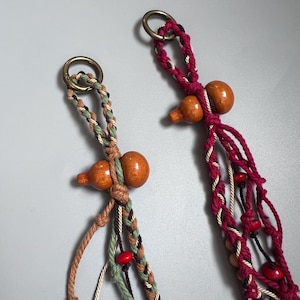 Puede incluir: Dos colgadores de plantas de macramé con cuentas de madera en forma de calabaza y anillos de latón. Un colgador presenta una combinación de colores marrón, verde y crema, mientras que el otro es burdeos con cuentas rojas. Estos colgadores decorativos están diseñados para sostener plantas en macetas.