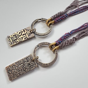 Puede incluir: Dos llaveros con colgantes rectangulares de metal con patrones y escritura en relieve, unidos a anillos circulares grabados con las palabras GOOD LUCK. Cada anillo se conecta a cordones trenzados en tonos gris, azul y rojo con nudos.