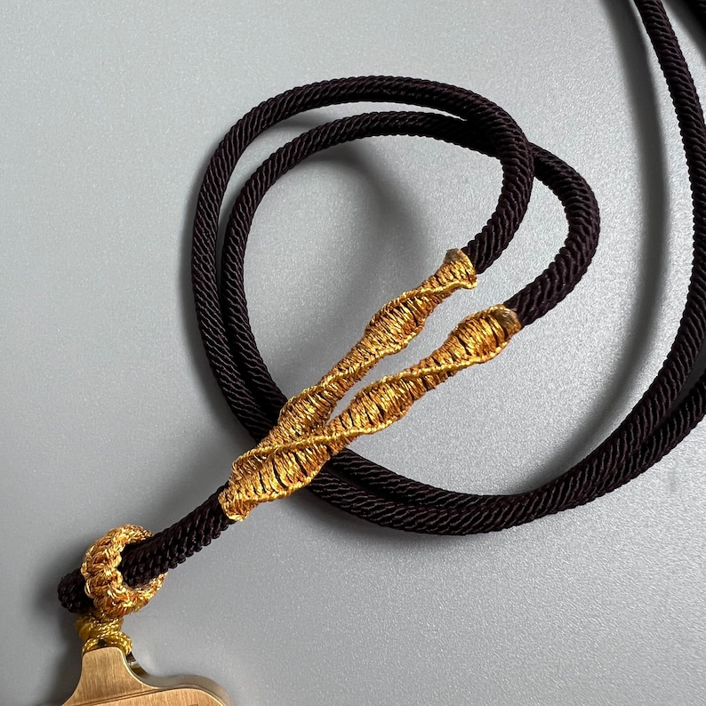 May include: A dark brown cord necklace featuring a decorative gold-colored metallic thread wrapping that secures the cord into a loop. The cord is braided and smooth, connecting to a metallic component at the bottom with a small gold-colored knot.