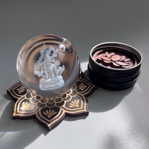 May include: A clear glass sphere with a detailed white figure inside, resting on a black and gold lotus flower-shaped base. Beside it is a small, open, black tin filled with wooden lotus flower cutouts. The items are on a gray surface.