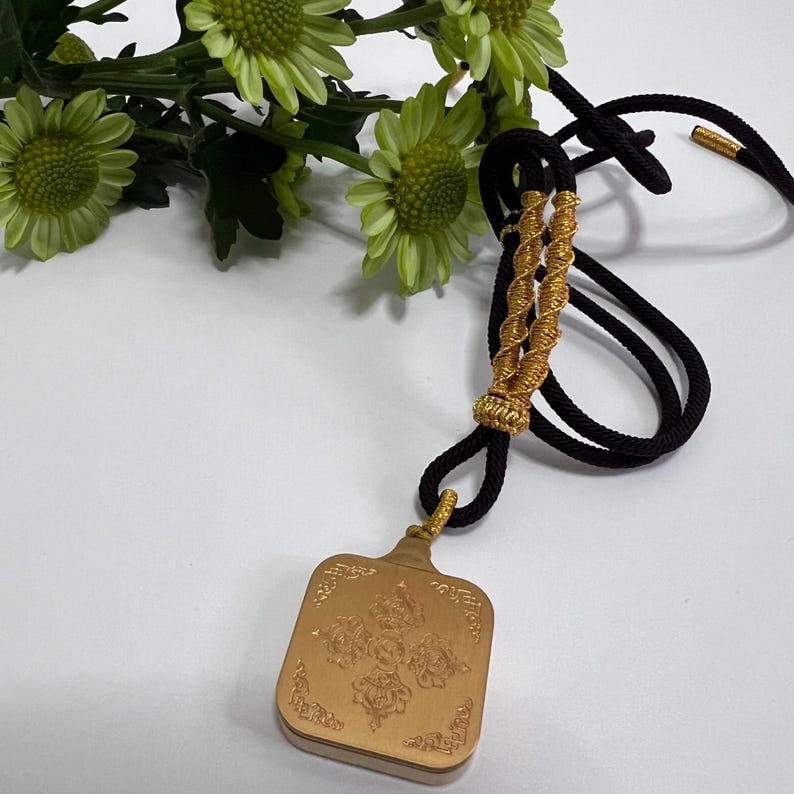 May include: A rectangular gold-toned pendant featuring an intricate embossed floral design on its surface. The pendant is suspended from a black cord that includes decorative gold-colored braided accents and gold-tipped ends.