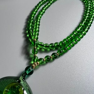 May include: A green glass beaded necklace with a green glass pendant. The beads are round and have a slight shimmer. The pendant is shaped like a leaf and has a gold accent.
