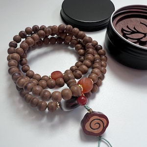 Könnte beinhalten: Eine hölzerne Gebetskette mit drei Strängen brauner Perlen. Das Mala hat rote und orangefarbene Edelsteinakzente und einen dekorativen Holzanhänger mit einem Spiralmuster. Ein schwarzer Deckel und ein kleiner Behälter befinden sich im Hintergrund.