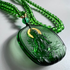 May include: A green glass necklace with a green glass pendant featuring a detailed carving of a deity. The pendant is attached to the necklace with a gold-colored chain.