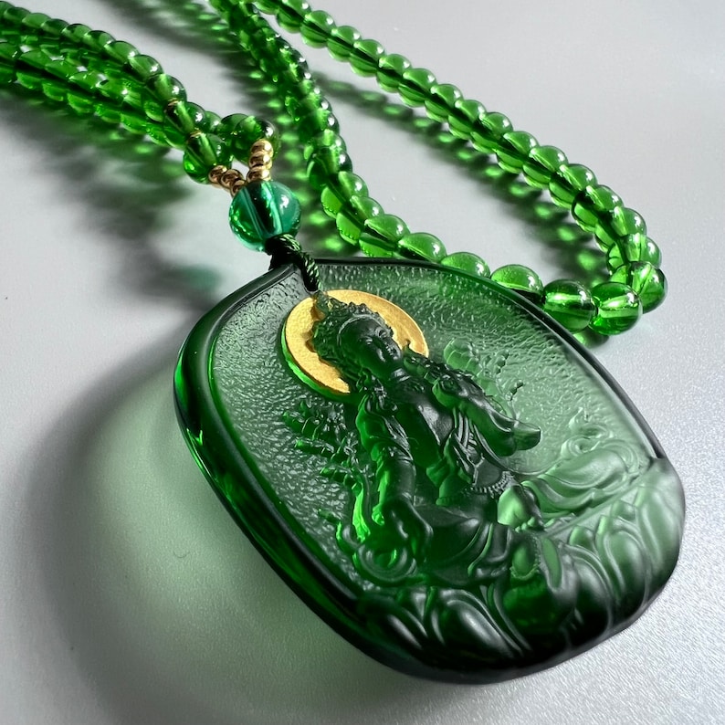 May include: A green glass necklace with a green glass pendant featuring a detailed carving of a deity. The pendant is attached to the necklace with a gold-colored chain.