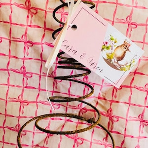 May include: A rusty metal spring with a pink tag attached by a white ribbon. The tag has the text "Cara & Wren" and a watercolor illustration of a bird with flowers. The background is a pink and white pattern of bows.