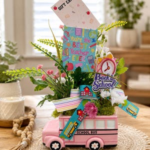 May include: A pink ceramic school bus planter filled with artificial greenery, pink flowers, and decorative signs. The bus features the text "SCHOOL BUS" and is adorned with ribbons, a "Back to School" sign, and a "You're the BEST Teacher EVER" card.