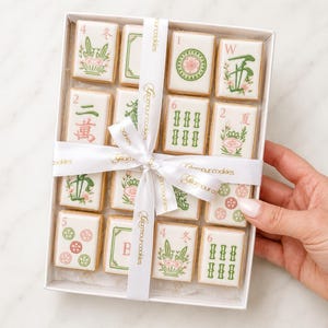 May include: A rectangular white box containing sixteen rectangular cookies decorated to resemble mahjong tiles. The cookies feature various green, red, and pink illustrations and characters. A white ribbon printed with the text Glamour cookies is tied around the box.
