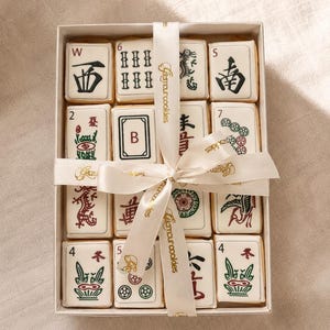 May include: A box of decorated cookies, each resembling a Mahjong tile. The cookies are white with various symbols and numbers in black, red, and green. A white ribbon with gold text wraps around the box.