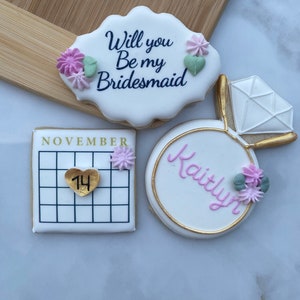 May include: Three decorated cookies. One cookie is white with the text "Will you Be my Bridesmaid" in black. Another cookie is white with a gold ring and a diamond. The third cookie is white with a calendar and the number 14 in a gold heart.